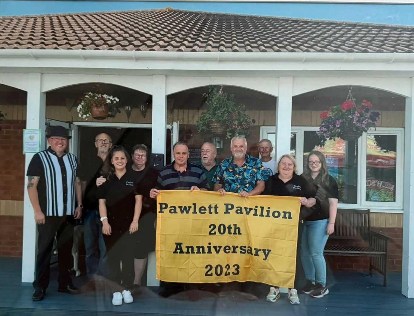 Volunteers and bar team outside the clubhouse holding a Pawlett Pavilion 20th Anniversary banner