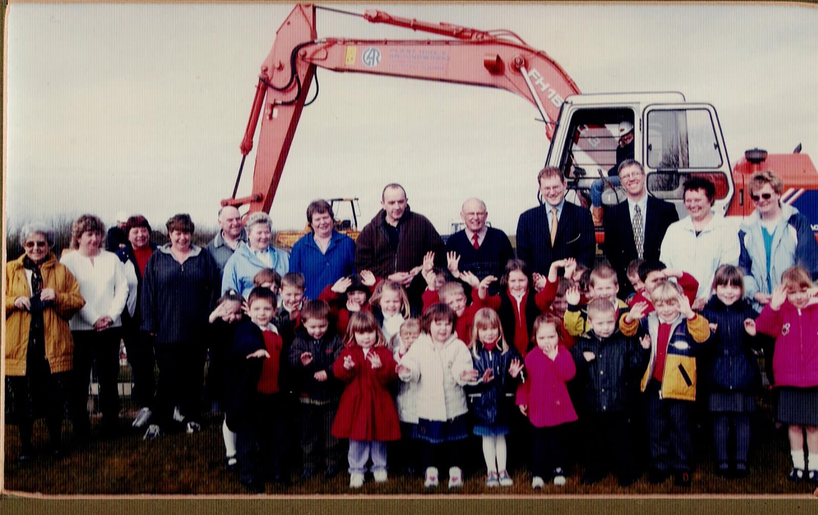 A crowd of waving schoolchildren and villagers in front of a digger at the first sod cutting